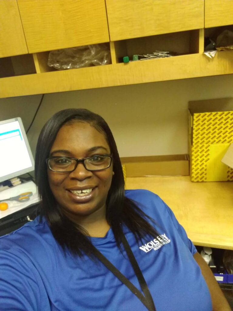 Smiling woman in blue shirt with "Wolsey" logo, seated at desk with computer, showcasing success in workplace, part of VIP Staffing success story series.