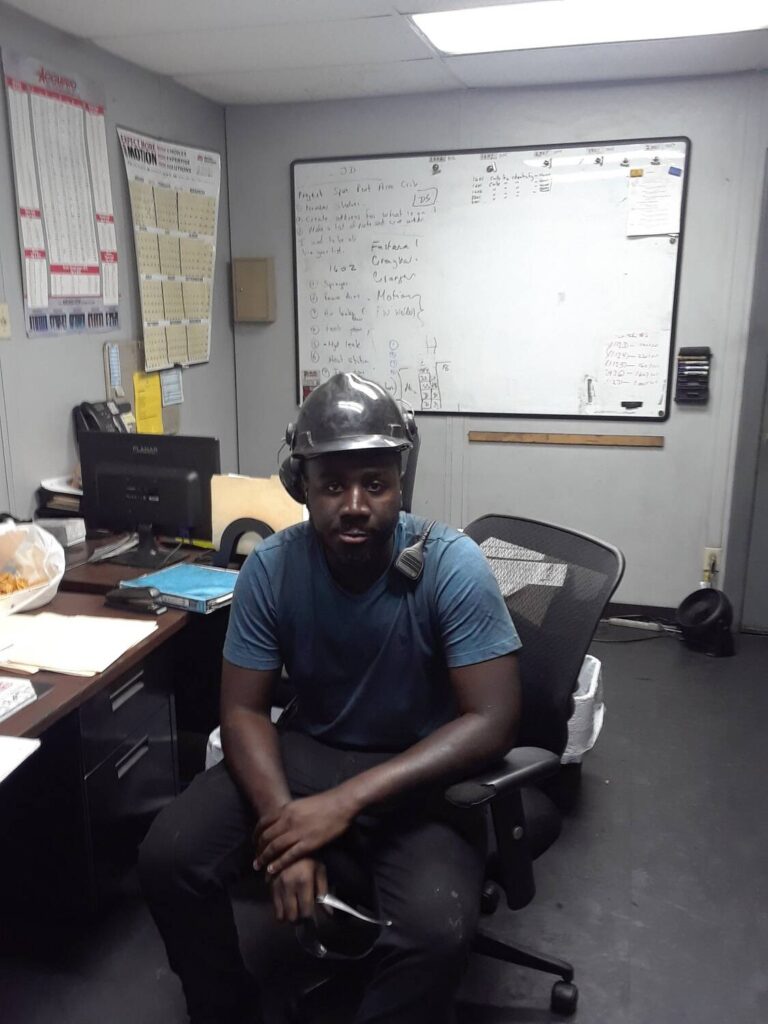Man sitting in an office chair wearing a hard hat and headset, surrounded by office equipment and a whiteboard with notes, representing success in the workplace through VIP Staffing.